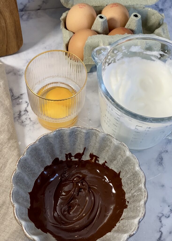 Sur une table les ingrédients nécessaires à la réalisation de la mousse au chocolat, dans différents bols. Le chocolat pâtissier est fondu et les blancs d’œufs battus en neige.
