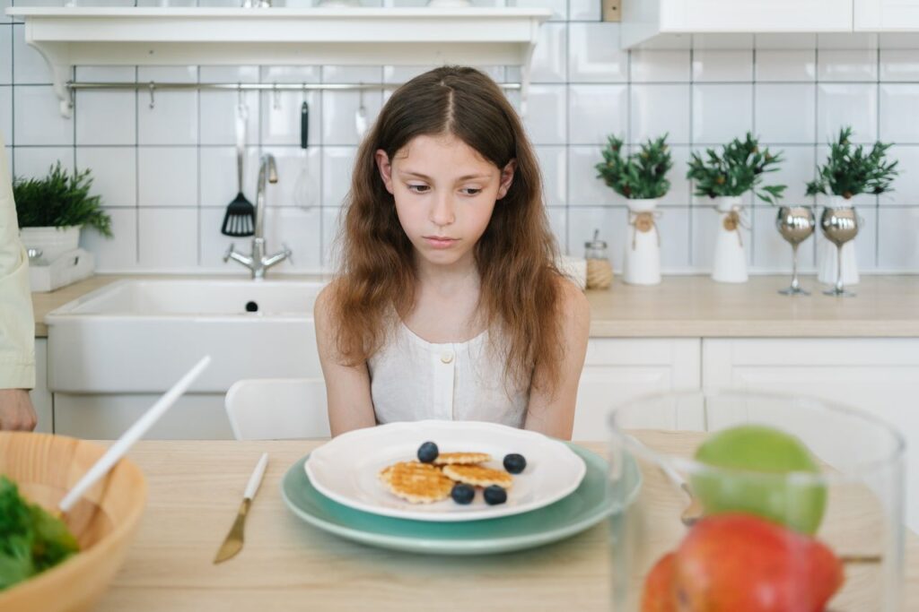 Petite fille dans une cuisine, assise devant une assiette de gaufres, avec une expression triste car elle ne peut pas manger de gluten